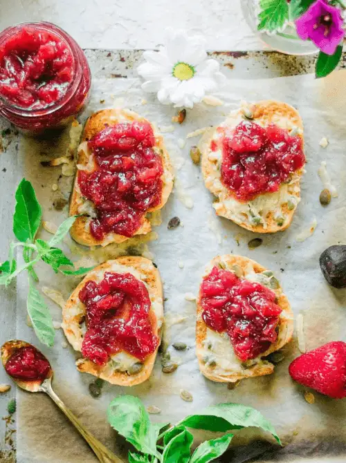 Toasted slices of crusty bread topped with melted cheese and a vibrant homemade strawberry jam, surrounded by fresh herbs, seeds, and flowers—these colorful bites make a charming and delicious addition to your Mother's Day tea party ideas.