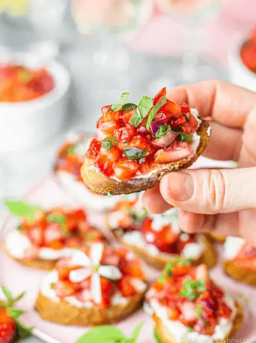 A hand holds a toasted crostini topped with whipped cream cheese, juicy diced strawberries, and fresh basil—these vibrant and sweet-savory bites are a fresh, colorful highlight for your Mother's Day tea party ideas.
