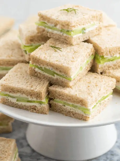 A stack of classic cucumber tea sandwiches made with soft wheat bread, creamy spread, and crisp cucumber slices is beautifully arranged on a white cake stand—an elegant and timeless choice for your Mother’s Day tea party ideas.S