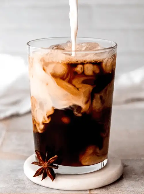 A tall glass of iced chai with milk swirling in rich patterns, garnished with a star anise and set on a marble coaster—an eye-catching and flavorful drink idea to elevate your Mother's Day tea party celebration.
