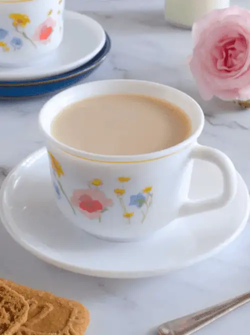 A delicate floral teacup filled with creamy tea sits on a matching saucer, surrounded by a pink rose, cookies, and a silver spoon—an elegant and charming setting perfect for Mother's Day tea party ideas.