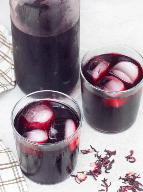 Two clear glasses filled with deep red hibiscus tea over ice sit beside a matching pitcher and scattered dried hibiscus petals—a bold, refreshing, and naturally floral option for your Mother's Day tea party ideas.