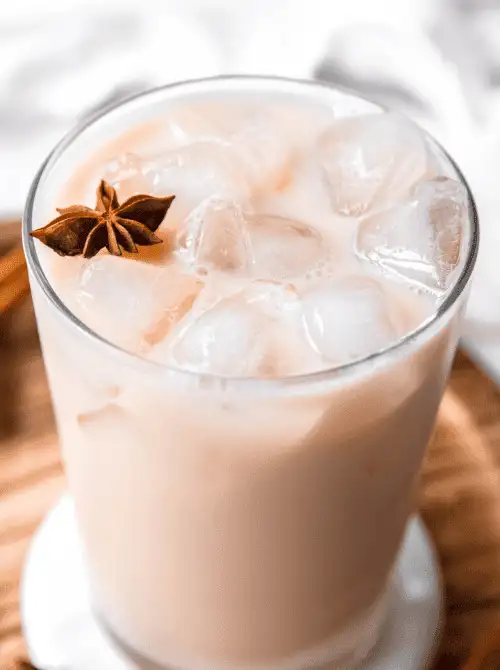 A refreshing glass of iced chai latte, filled with ice cubes and garnished with a star anise pod, rests on a wooden tray—an elegant and cooling twist to consider for your Mother's Day tea party ideas.