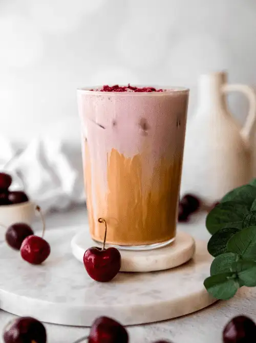 A layered iced cherry latte in a tall glass, topped with crushed cherries and surrounded by fresh fruit on a marble tray—this vibrant and fruity beverage is a sweet and whimsical choice for Mother's Day tea party ideas.