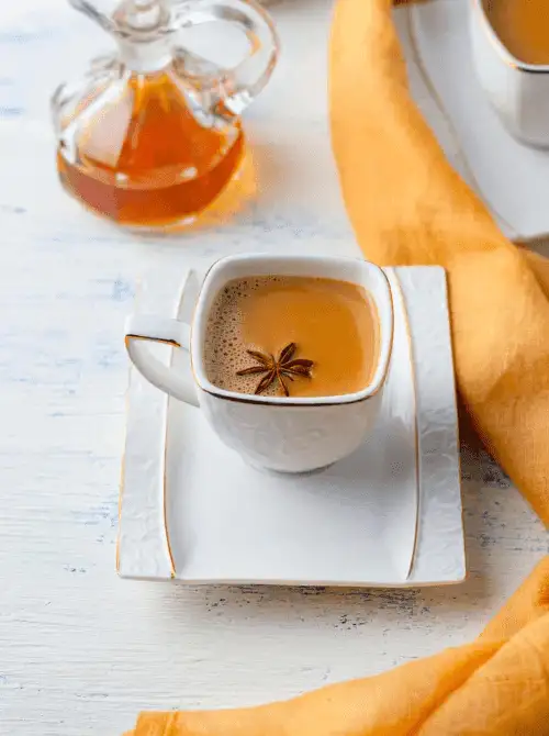 A sophisticated white teacup filled with spiced tea and topped with a star anise floats on a matching saucer set against a warm yellow cloth and honey jar—perfect for elegant and aromatic Mother's Day tea party ideas.