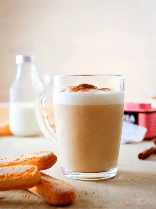 A cozy glass mug filled with a frothy latte, topped with a sprinkle of cinnamon, sits on a light wooden table surrounded by ladyfinger cookies and a small milk bottle in the background—perfect for cozy Mother's Day tea party ideas.