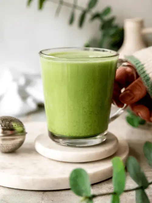 A glass mug of vibrant green matcha latte sits on a marble coaster, surrounded by fresh greenery and cozy textures—an earthy and calming option to include in your Mother's Day tea party ideas.