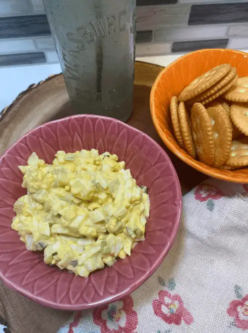 A pink bowl filled with creamy Pickled Onion Egg Salad, served on a wooden tray beside buttery crackers in an orange bowl, perfect for a budget-friendly brunch or Mother's Day tea party.