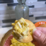 A close-up of a hand holding a round cracker topped with creamy egg salad, with a bowl of egg salad and crackers blurred in the background.