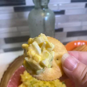 A close-up of a hand holding a round cracker topped with creamy egg salad, with a bowl of egg salad and crackers blurred in the background.