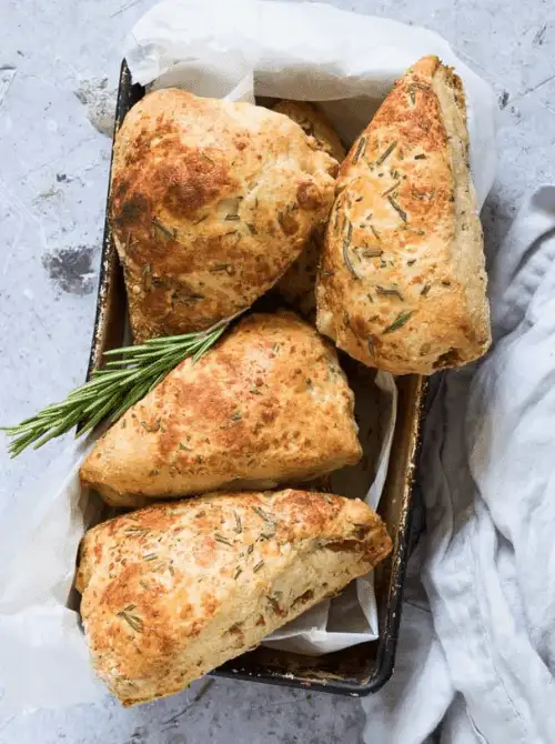 Flaky rosemary scones with golden crusts nestled in a parchment-lined tray, garnished with fresh rosemary sprigs—an earthy, savory option for elegant Mother's Day tea party ideas.
