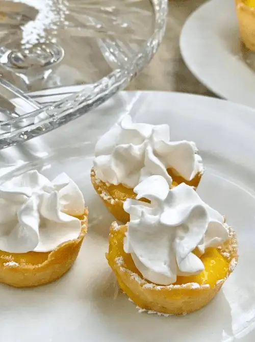 Mini lemon tarts topped with fluffy whipped cream and a dusting of powdered sugar, served on elegant white china—bright and zesty treats perfect for springtime Mother's Day tea party ideas.