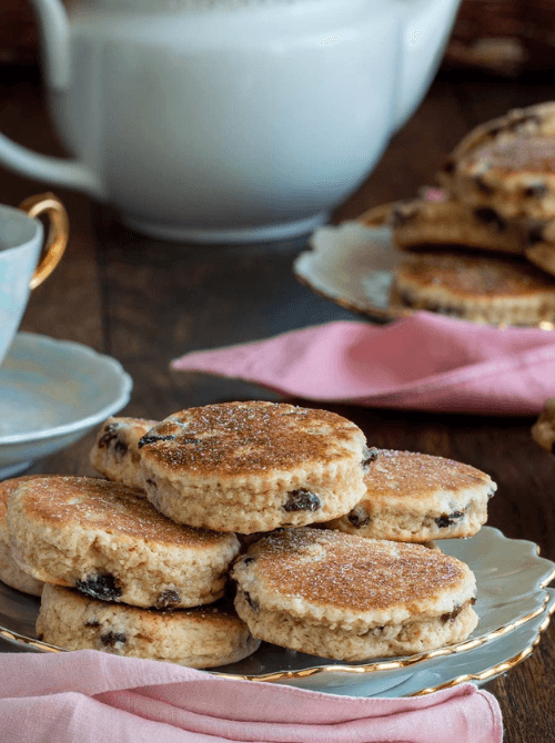 A vintage tea setting with golden-edged china plates stacked with traditional Welsh cakes studded with currants, set beside pink napkins and a teapot—perfect for cozy and charming Mother's Day tea party ideas.
