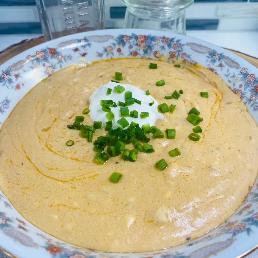 Warm gouda, tahini, and cream cheese dip served in a floral-patterned dish, topped with a dollop of sour cream, chopped green onions, and a swirl of olive oil.
