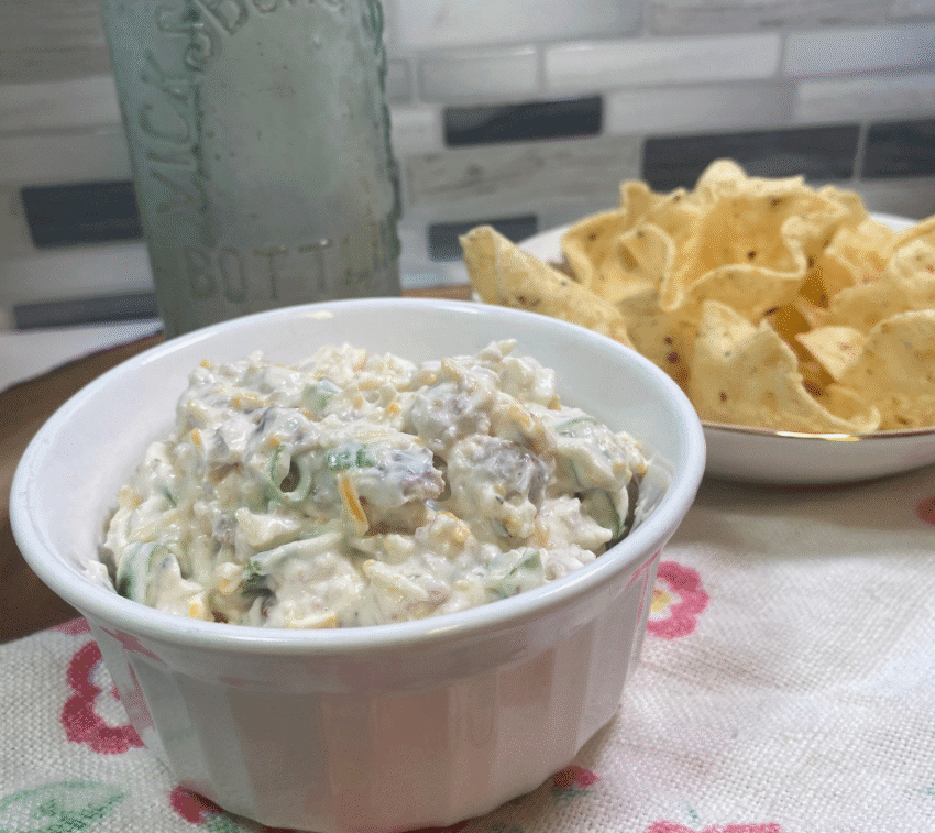 Cheeseburger Ranch Dip served in a white bowl, filled with chopped burger, shredded cheese, green onions, sour cream, and mayo. A creamy, no-cook appetizer shown with tortilla chips in the background—perfect for parties, potlucks, or budget-friendly snacking.