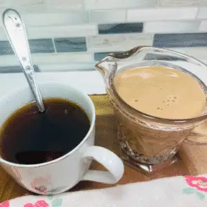 Homemade chocolate caramel coffee creamer in a glass pitcher next to a cup of black coffee with a silver spoon, styled on a wooden tray with a tiled backsplash.