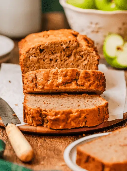 Sliced apple bread loaf on a wooden board with fresh green apples in the background—a cozy fall bread recipe and a tasty idea for what to make with apples.