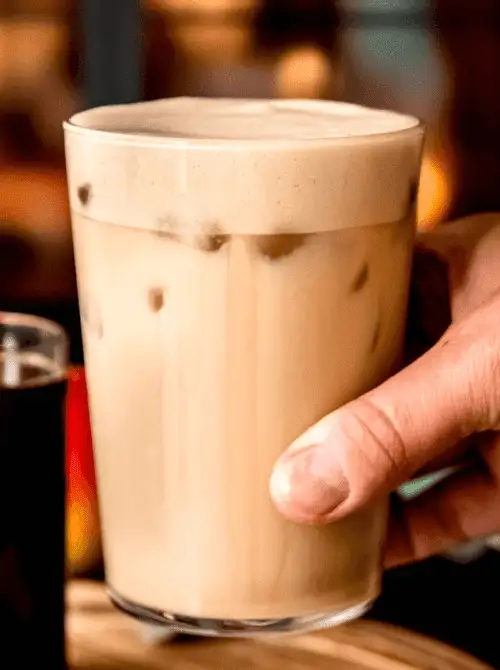Hand holding an iced apple crisp shaken espresso in a tall glass with creamy foam—refreshing and flavorful, this seasonal drink is one of the easiest fall recipes and trendiest things to make with apples.