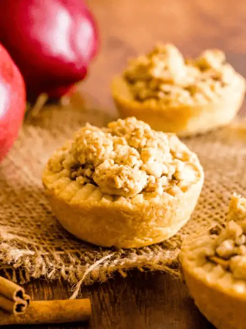 Mini apple crumble tartlets on rustic burlap with fresh apples and cinnamon—bite-sized fall desserts and adorable things to make with apples for cozy gatherings.