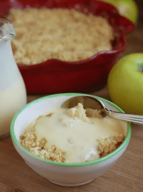 Warm apple crumble served with custard in a bowl, with a baking dish and green apples in the background—classic fall dessert comfort and one of the most nostalgic things to make with apples.