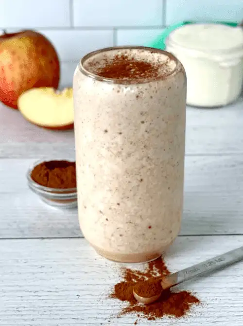 Creamy apple cinnamon smoothie in a tall glass, topped with ground cinnamon and surrounded by fresh apples and spices—an easy fall recipe and a refreshing twist on things to make with apples.