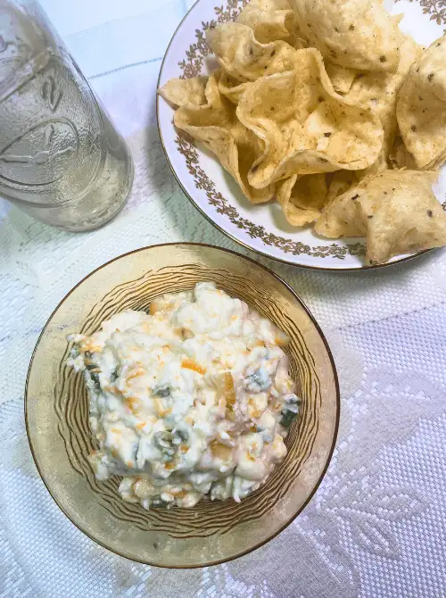 Bowl of creamy apricot jalapeño popper dip with visible green onions and shredded cheese, served with a side plate of tortilla chips on a vintage tablecloth. Perfect for game day snacks or easy party appetizers.