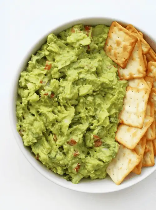 A white bowl filled with chunky guacamole and square crackers—one of the easy game day dips perfect for serving a crowd.