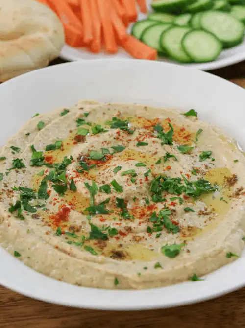 A bowl of homemade hummus garnished with olive oil, paprika, and fresh parsley, served with carrot sticks, cucumber slices, and pita—perfect for budget-friendly appetizers, easy snacks to feed a crowd, or a healthy football party dip.