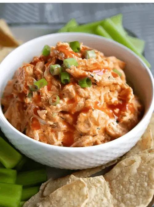 Buffalo chicken dip topped with green onions and hot sauce, served in a white bowl with celery sticks and tortilla chips—easy tailgate food on a budget for a crowd.