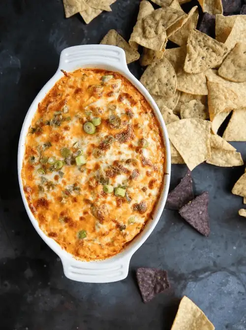 Hot cheesy dip in a white baking dish topped with green onions, surrounded by assorted tortilla chips — a melty, crave-worthy addition to your lineup of easy game day dips.