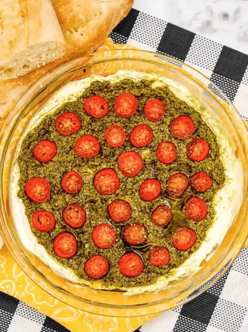 A glass dish filled with creamy cheese layered with vibrant green pesto and topped with halved cherry tomatoes, served alongside crusty bread—ideal for football party snacks, easy appetizers, and game day food on a budget.