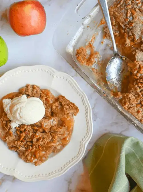 Serving of warm apple crumble topped with vanilla ice cream on a white plate beside a baking dish—one of the best fall desserts and a classic choice when looking for cozy things to make with apples.