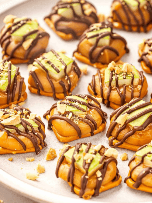 Things to make with apples for simple fall treats – caramel apple pretzel bites topped with fresh green apple slices, melted caramel, crushed nuts, and chocolate drizzle on a serving tray.