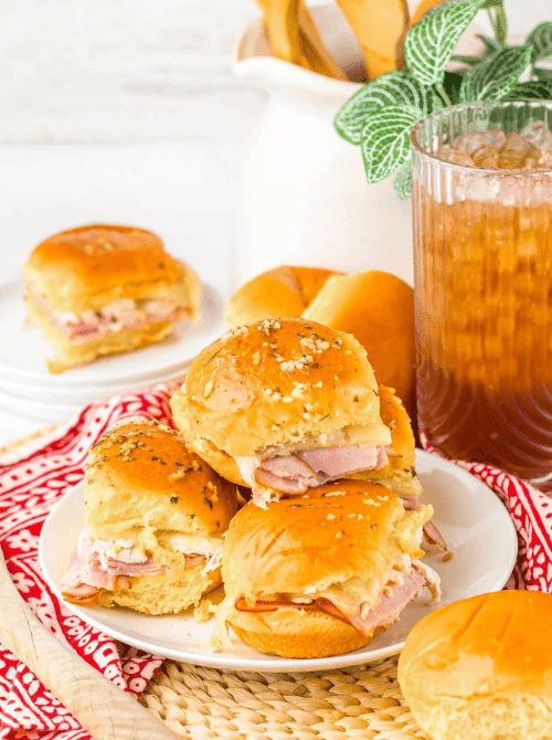 Stacked ham and cheese sliders on golden buns with buttery topping, served on a white plate—easy tailgate food on a budget that’s perfect for a crowd.
