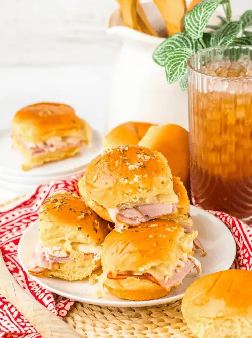 Stacked ham and cheese sliders on golden buns with buttery topping, served on a white plate—easy tailgate food on a budget that’s perfect for a crowd.