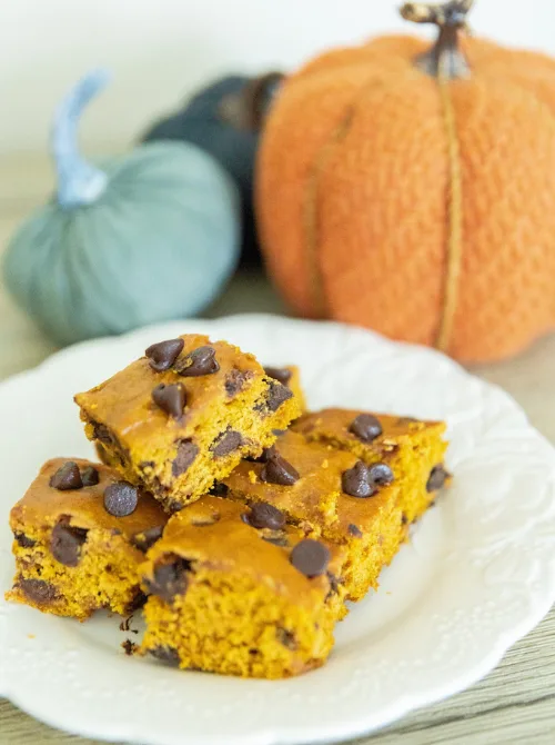 Pumpkin chocolate chip bars stacked on a white plate with cozy fall decor in the background—an easy pumpkin dessert and a delicious way to use leftover pumpkin puree.