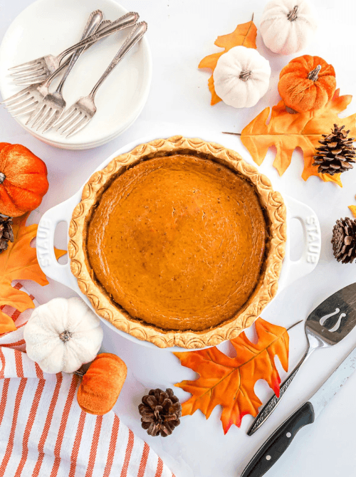 Whole pumpkin pie in a white baking dish surrounded by mini pumpkins, pinecones, and fall leaves—perfect for fall gatherings and a festive way to use leftover pumpkin puree in an easy pumpkin pie.