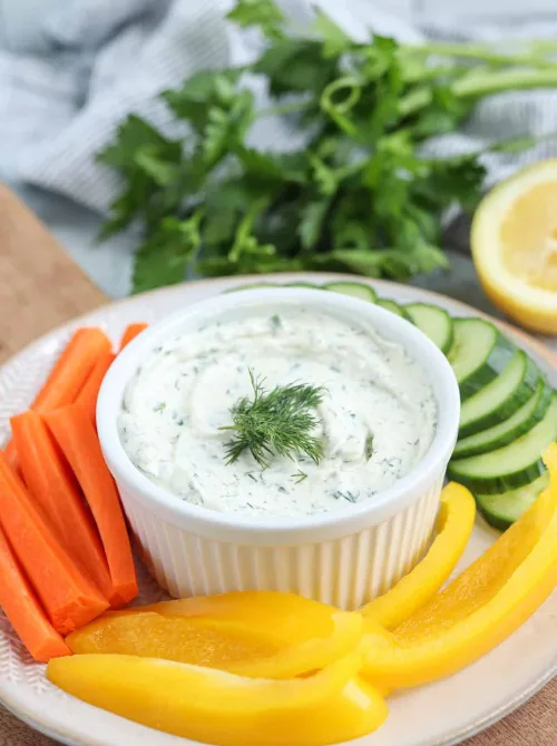 Creamy herb dip in a white ramekin, garnished with dill and surrounded by fresh carrot sticks, yellow bell peppers, and cucumber slices — a light and refreshing option for easy game day dips.