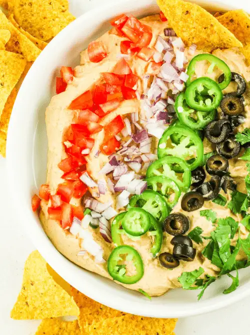 Colorful layered dip topped with diced tomatoes, red onions, jalapeños, black olives, and cilantro in a white bowl, surrounded by tortilla chips — a bold and fresh option for easy game day dips.