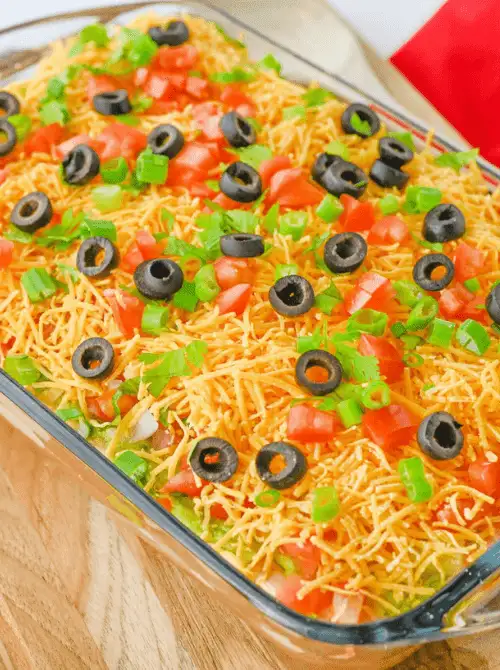 Glass dish filled with layered taco dip topped with shredded cheddar cheese, diced tomatoes, sliced olives, and green onions — a vibrant and budget-friendly option for easy game day dips.