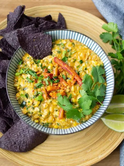 A colorful bowl of Mexican street corn dip garnished with crispy tortilla strips, fresh cilantro, and green onions, served with blue corn chips—perfect for football party snacks, tailgate appetizers, and budget-friendly game day recipes.