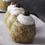 Round apple desserts coated in finely chopped nuts, topped with a swirl of whipped cream and a cherry, arranged neatly on a rectangular serving dish for an elegant presentation.