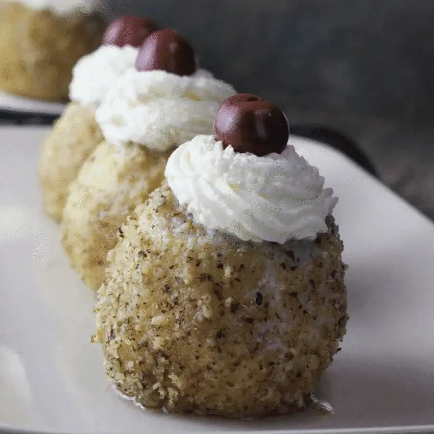 Round apple desserts coated in finely chopped nuts, topped with a swirl of whipped cream and a cherry, arranged neatly on a rectangular serving dish for an elegant presentation.