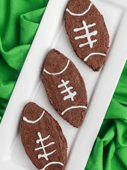 Chocolate sugar cookies decorated like footballs with white icing, displayed on a white platter—an easy and affordable option for football party snacks on a budget that adds festive flair to any game day dessert spread.