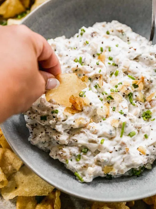 A hand dipping a potato chip into a creamy onion and chive dip—one of the best easy game day dips to pair with crunchy snacks.