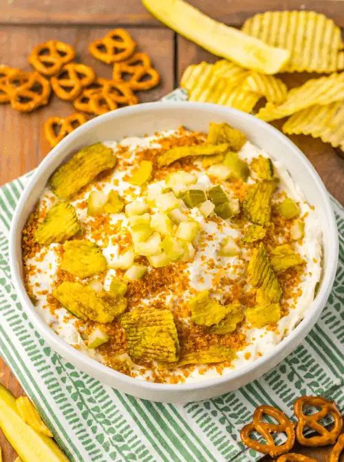 A white bowl filled with creamy pickle dip topped with chopped pickles, crushed chips, and pretzels, surrounded by snacks—perfect for serving easy game day dips with bold flavor.