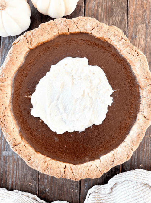 Overhead view of a rustic pumpkin pie topped with a generous swirl of whipped cream, set on a wooden table with white mini pumpkins—a cozy, homemade way to use leftover pumpkin puree for an easy fall dessert.