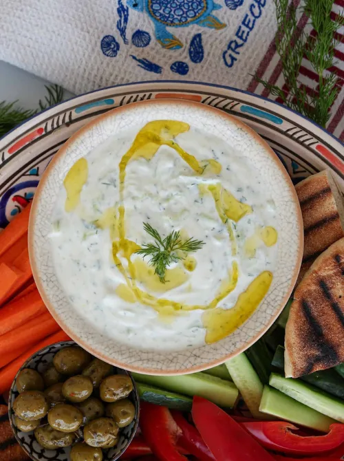 A bowl of creamy tzatziki drizzled with olive oil and topped with fresh dill, surrounded by a colorful spread of Mediterranean veggies, pita bread, and olives—perfect for a healthy game day snack or football finger foods with a fresh twist.