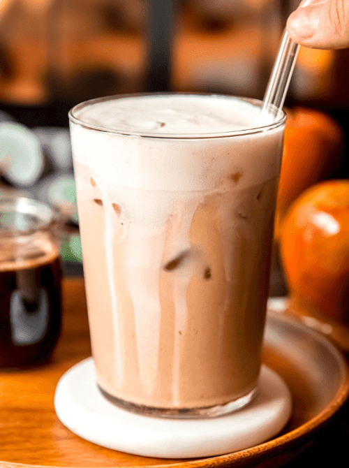 Iced apple crisp latte in a tall glass with creamy foam and a straw—smooth, spiced, and a cozy coffeehouse-inspired drink that’s perfect for easy fall recipes and delicious things to make with apples.