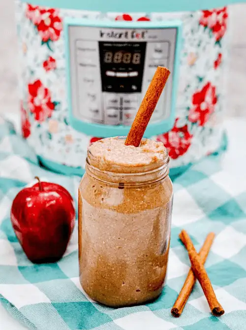 Homemade cinnamon applesauce in a mason jar with a cinnamon stick, placed in front of a floral Instant Pot—an easy fall recipe and a cozy, nostalgic way to use up apples.